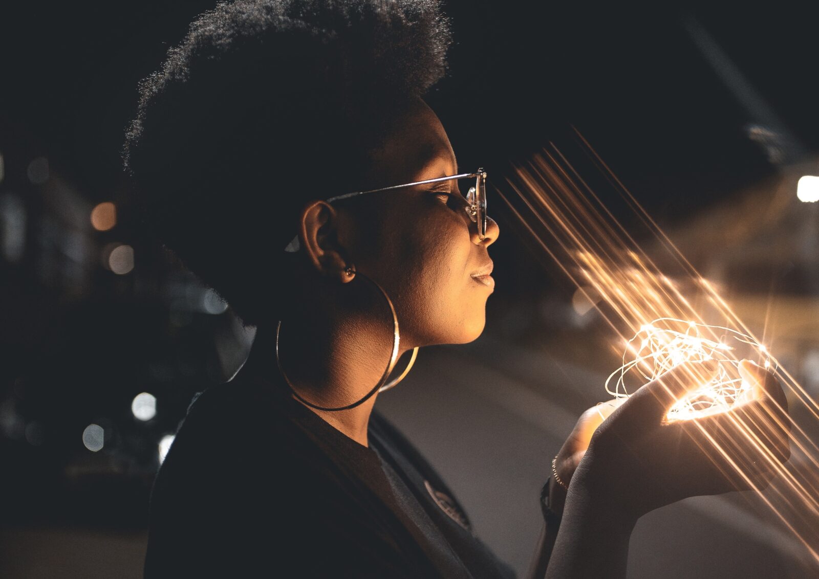 Black woman with glasses and hoop earrings standing in the dark holding a string of lit fairy lights with a peaceful look on her face