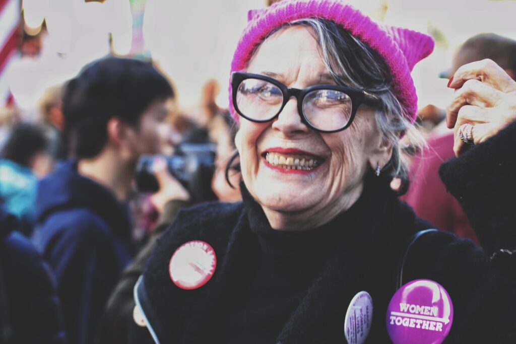 Woman smiling with glasses and a pink hat at an event with people surrounding her