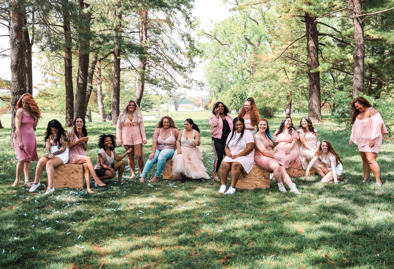 Group of diverse women all lined up on a grassy area in mostly pink clothing, smiling, surrounded by sunlight and trees