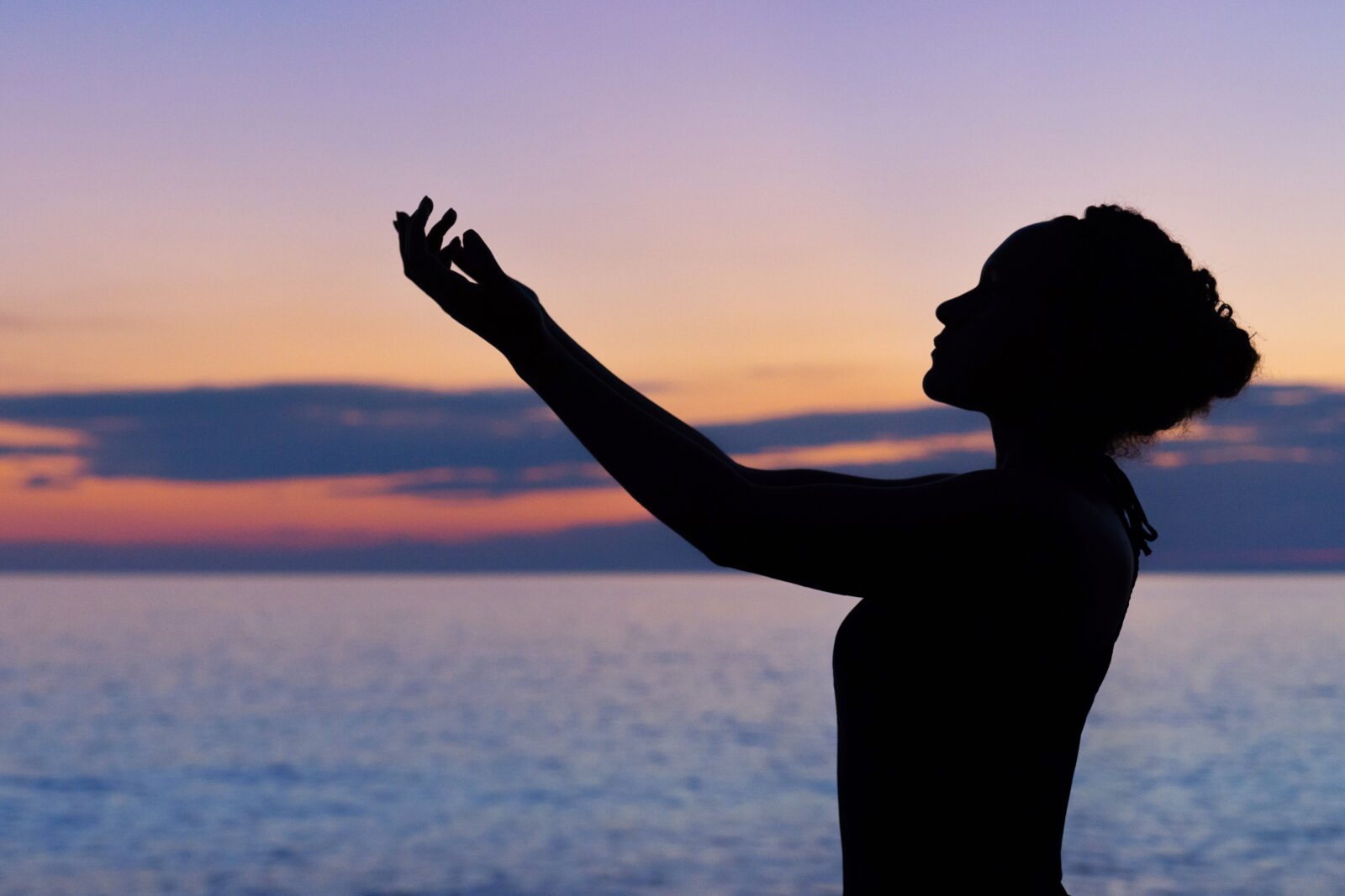 Silhouette of woman, arms reaching out and up in front of her lit by a pastel sky