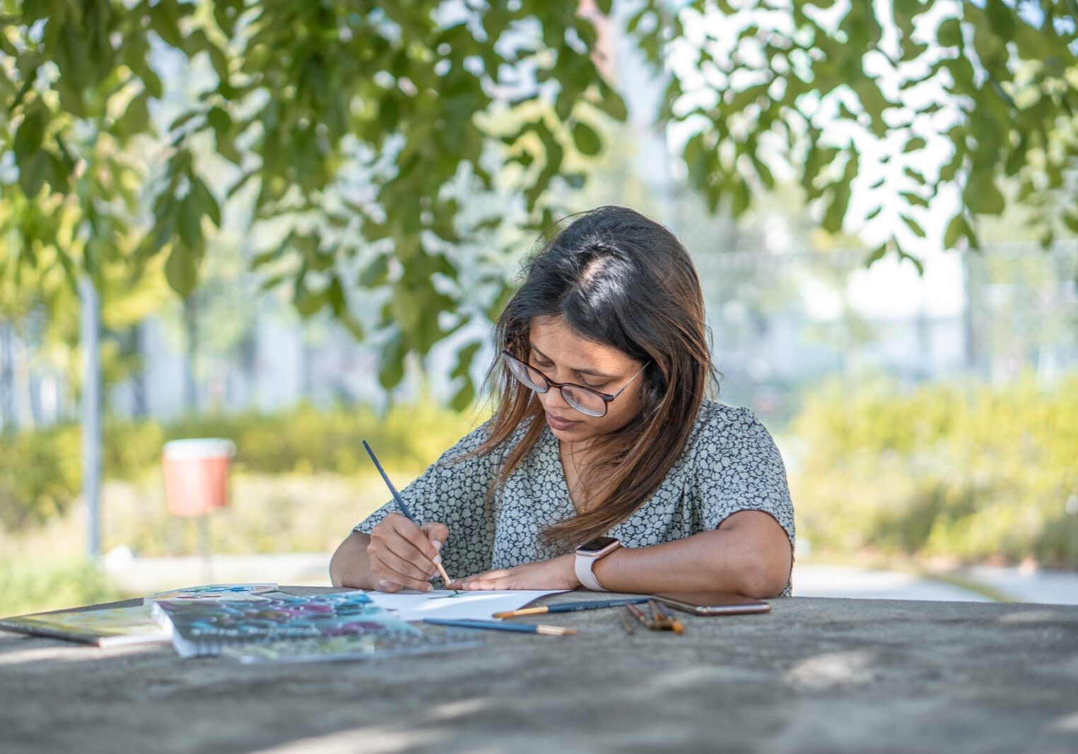 Woman with long hair and glasses sitting at a table in the shade under a tree on a sunny day painting.