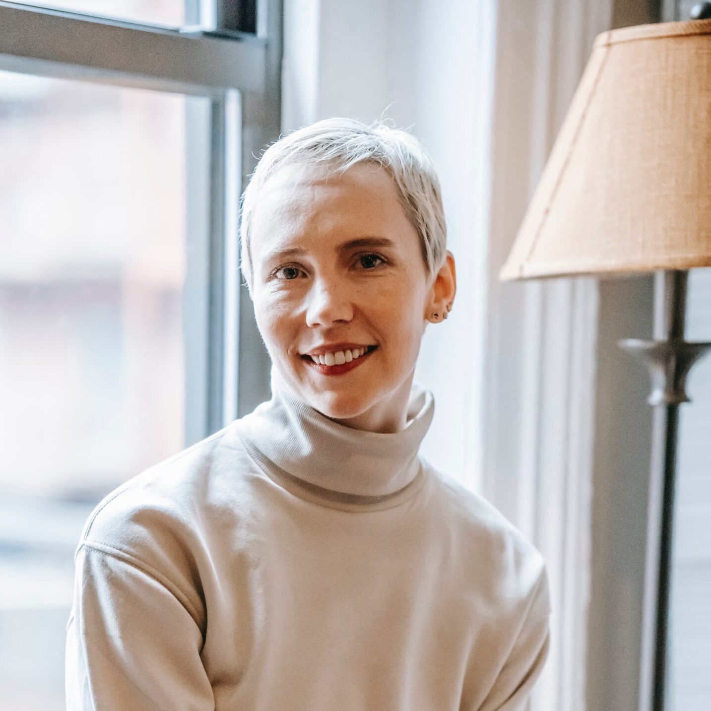 Woman with short cropped gray hair sitting next to a lamp in front of a window wearing a beige turtleneck and smiling at the camera