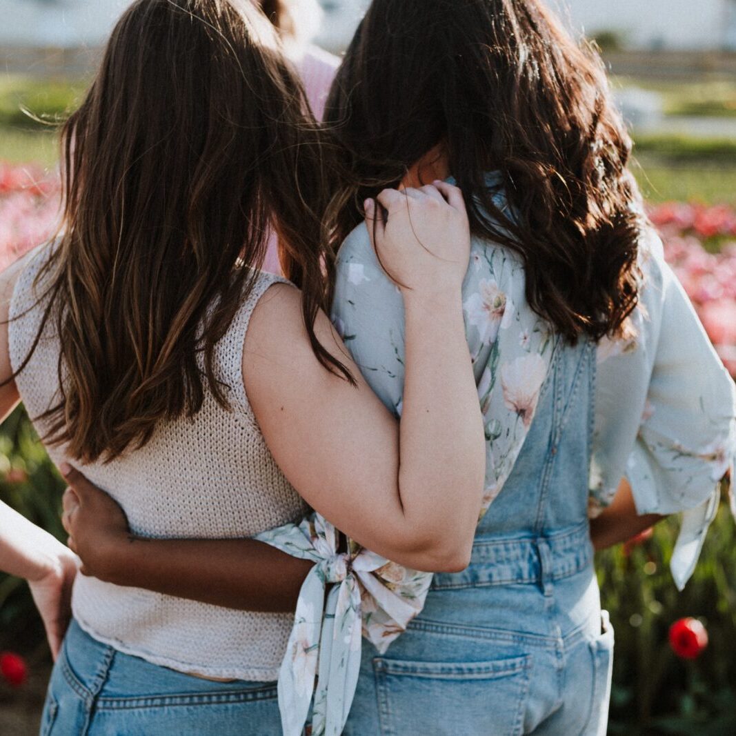 Two women standing facing away from the camera, in the sun and in front of a field of mostly red and pink flowers, arms closely around each other.