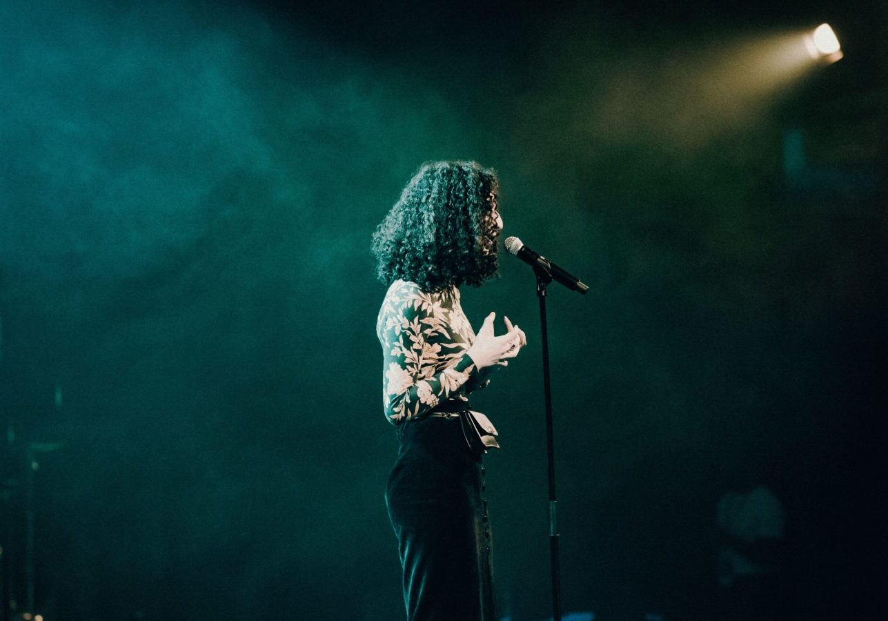 Woman standing on a stage in a dark room speaking into a mic with hands gesturing as she speaks